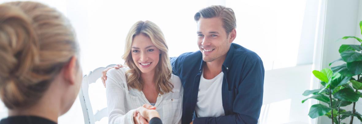 Real estate agent with couple shaking hands closing a deal. There is a digital tablet on the table. Couple are casually dressed. They are looking happy and smiling. Over the shoulder view inside a home.