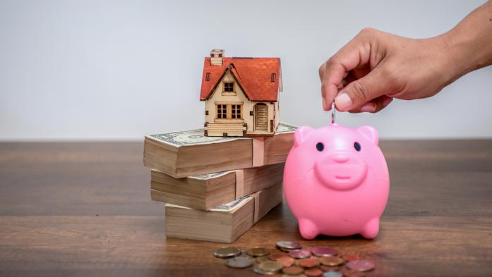 Toy home on top of money beside a piggy bank